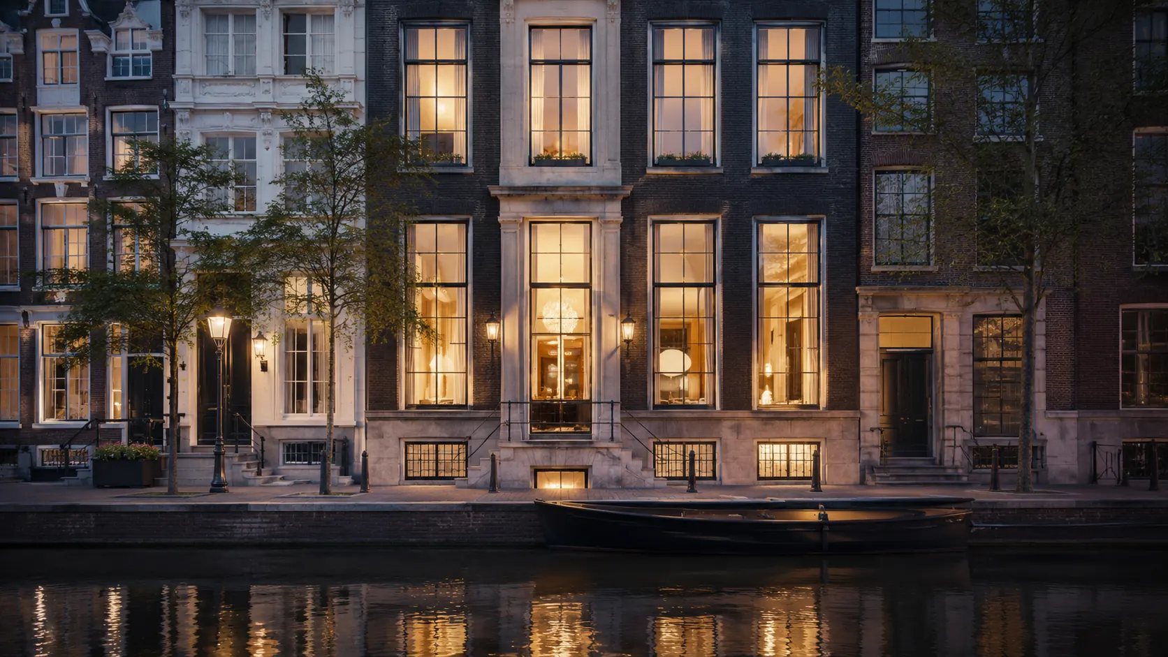 A row of Amsterdam canal houses at night, tall sash windows lit warm from within, reflected in the water.